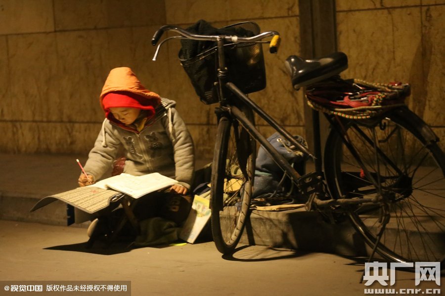 鄭州中原路京廣鐵路大橋下涵洞的馬路牙子邊，一位戴著紅色辣椒帽的小男孩趴在一個(gè)小馬扎上，上面鋪著一張車墊，在微弱的燈光下寫作業(yè)